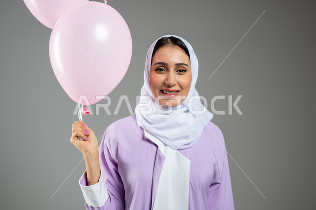 النظر إلى الكاميرا بتعابير الثقة والسرور, مفهوم الأناقة والاهتمام بالمظهر الخارجي, بورتريه مقرب لشابة عربية خليجية سعودية مبتسمة ترتدي العباءة الملونة والحجاب تمسك بيدها مجموعة من البلالين الوردية, خلفية رمادية, محتوى حساس, استخدام حساس