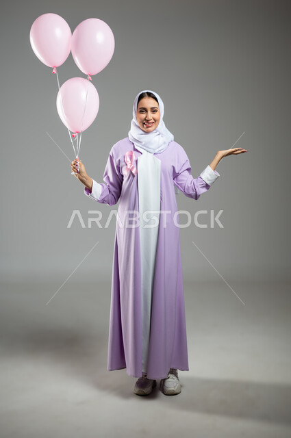 حمل عنصر معين, الحملات التوعوية ضد الأمراض الخبيثة, اليوم العالمي للكشف المبكر عن سرطان الثدي, بورتريه لإمرأة عربية خليجية سعودية مبتسمة ترتدي العباءة تضع الشريط الوردي وتمسك بيدها البلالين, خلفية رمادية, صورة الجسم بالكامل, محتوى حساس, استخدام حساس