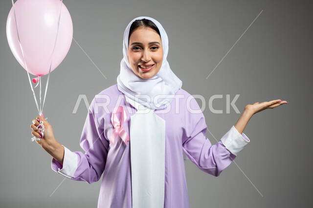 الإشارة إلى شيء ما وحمل عنصر معين, الحملات التوعوية ضد الأمراض الخبيثة, اليوم العالمي للكشف المبكر عن سرطان الثدي, بورتريه مقرب لإمرأة عربية خليجية سعودية مبتسمة ترتدي العباءة تضع الشريط الوردي وتمسك بيدها البلالين, خلفية رمادية, محتوى حساس, استخدام حساس