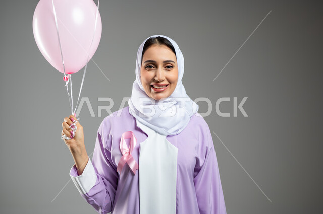 اليوم العالمي للكشف المبكر عن سرطان الثدي, الحملات التوعوية ضد الأمراض الخبيثة, بورتريه مقرب لإمرأة عربية خليجية سعودية مبتسمة محجبة ترتدي العباءة تضع الشريط الوردي وتمسك بيدها مجموعة من البلالين وتنظر إلى الكاميرا بقوة, خلفية رمادية, محتوى حساس, استخدام حساس