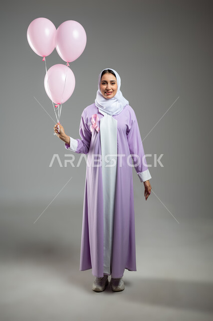 الحملات التوعوية ضد الأمراض الخبيثة, بورتريه لإمرأة عربية خليجية سعودية مبتسمة محجبة ترتدي العباءة تضع الشريط الوردي وتمسك بيدها مجموعة من البلالين, اليوم العالمي للكشف المبكر عن سرطان الثدي, صورة الجسم بالطول الكامل, خلفية رمادية, محتوى حساس, استخدام حساس