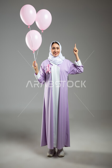 اليوم العالمي للكشف المبكر عن الأمراض الخبيثة, بورتريه لشابة عربية خليجية سعودية ترتدي العباءة تضع الشريط الوردي وتشير بالإصبع السبابة إلى الأعلى, تقديم الدعم ضد سرطان الثدي, صورة الجسم بالطول الكامل, خلفية رمادية, محتوى حساس, استخدام حساس