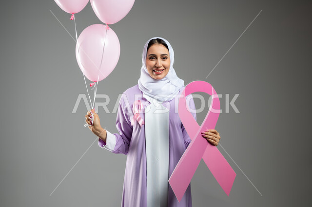 النظر إلى الكاميرا بتعابير القوة, اليوم العالمي للتوعية والكشف المبكر عن سرطان الثدي, بورتريه مقرب لإمرأة عربية خليجية سعودية مبتسمة ترتدي العباءة تمسك بيدها البلالين الوردية وشعار الحملة العالمية, تحدي الأمراض الخبيثة, خلفية رمادية, محتوى حساس, استخدام حساس