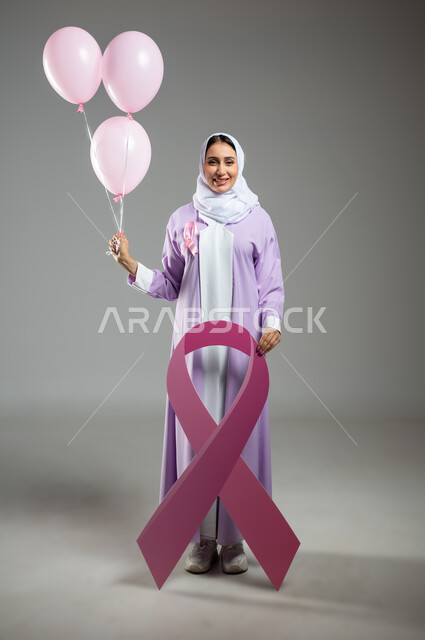الوقوف بقوة والنظر إلى الكاميرا, اليوم العالمي للتوعية والكشف المبكر عن سرطان الثدي, بورتريه لإمرأة عربية خليجية سعودية مبتسمة ترتدي العباءة تمسك بيدها البلالين الوردية, تحدي الأمراض الخبيثة, صورة الجسم بالطول الكامل, خلفية رمادية, محتوى حساس, استخدام حساس