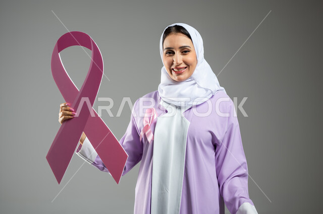 تقديم المساعدة والدعم من خلال الكشف المبكر عن سرطان الثدي, بورتريه مقرب لشابة عربية خليجية سعودية محجبة مبتسمة ترتدي العباءة الملونة تمسك بيدها شعار الحملة العالمية الوردي لتحدي الأمراض الخبيثة, خلفية رمادية, محتوى حساس, استخدام حساس