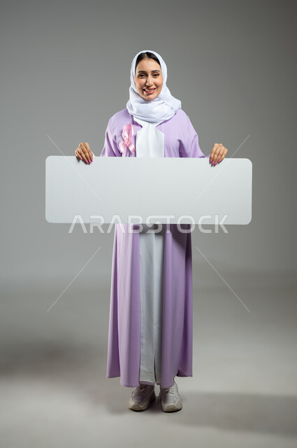 النظر إلى الكاميرا بإيماءات السرور, لوحات إعلانية للتوعية ضد الأمراض الخبيثة, بورتريه لشابة عربية خليجية سعودية ترتدي العباءة وتضع شعار الحملة العالمية لسرطان الثدي وتمسك بيدها موك أب مستطيل فارغ, صورة الجسم بالكامل, خلفية رمادية, محتوى حساس, استخدام حساس
