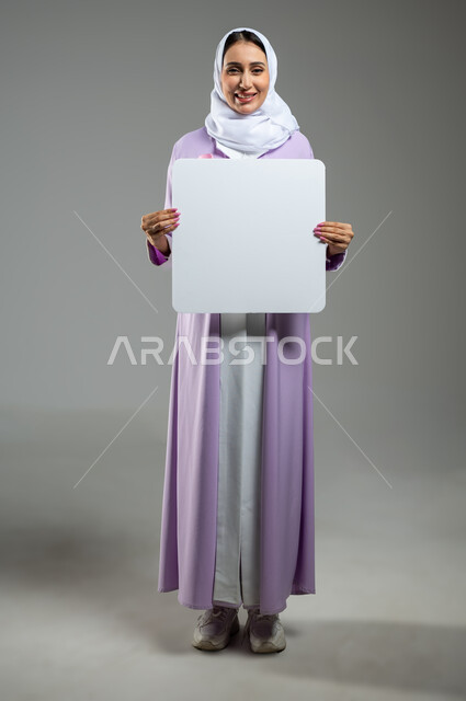 الوقوف بثقة والنظر إلى الكاميرا بابتسامة, لوحات إعلانية فارغة, بورتريه لشابة عربية خليجية سعودية محجبة ترتدي العباءة الملونة تمسك بيدها موك أب مربع أبيض بإيماءات البشاشة والسرور, صورة الجسم بالطول الكامل, خلفية رمادية, محتوى حساس, استخدام حساس