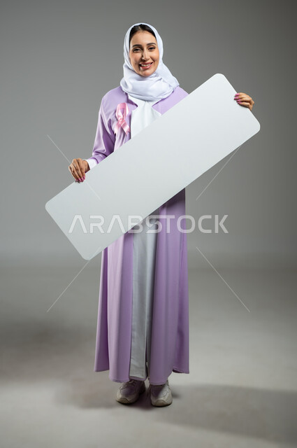 A blank white rectangular mockup display, the logo of the Global Campaign to Challenge Malignant Diseases, a portrait of a Saudi Gulf Arab young woman wearing a colorful abaya, wearing a pink ribbon and holding in her hand a billboard for breast cancer awareness, full length body image, gray background, sensitive content, sensitive use