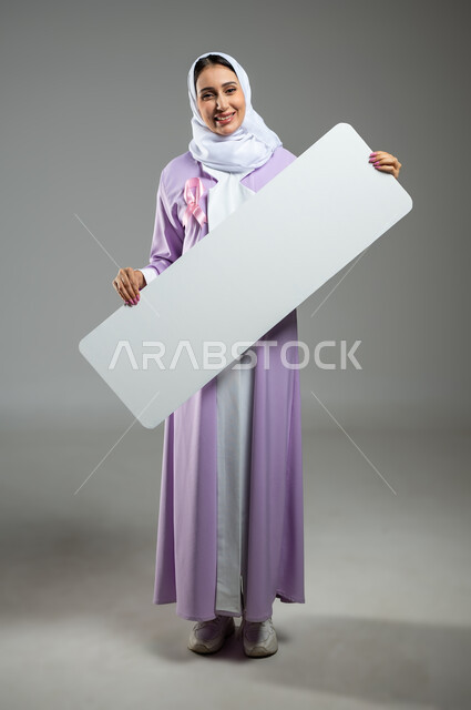 شعار الحملة العالمية لتحدي الأمراض الخبيثة, لوحات إعلانية للتوعية بسرطان الثدي, بورتريه لشابة عربية خليجية سعودية مبتسمة ترتدي العباءة تضع شريط وردي وتمسك بيدها موك أب مستطيل أبيض فارغ, صورة الجسم بالطول الكامل, خلفية رمادية, محتوى حساس, استخدام حساس