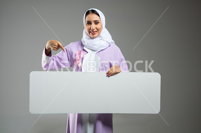 شعار اليوم العالمي للتوعيه بالكشف المبكر لسرطان الثدي، الحملة التوعويه بالامراض الخبيثه، بورتريه لامرأة عربية خليجية سعودية مبتسمة محجبة ترتدي العباءة  مع الشريط الوردي تشير بإصبع السبابة إلى موك اب مستطيل ابيض فارغ، خلفية رمادية، محتوى حساس، استخدام حساس