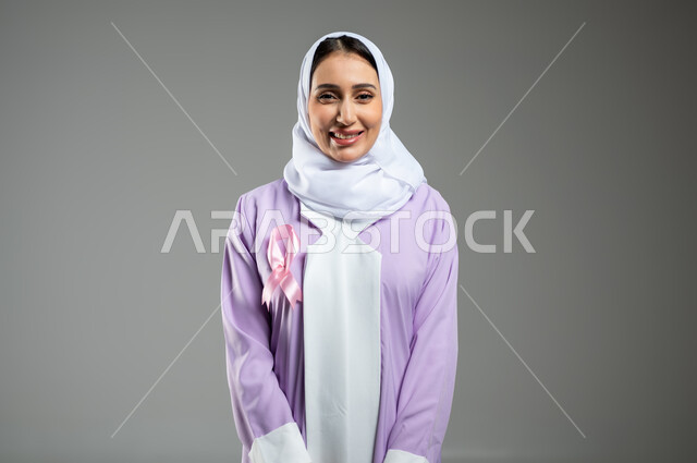 شعار اليوم العالمي للتوعيه بالكشف المبكر لسرطان الثدي، الحملة التوعويه بالامراض الخبيثه، بورتريه لامرأة عربية خليجية سعودية مبتسمة محجبة ترتدي العباءة مع الشريط الوردي تقف باستقامة، خلفية رمادية، محتوى حساس، استخدام حساس