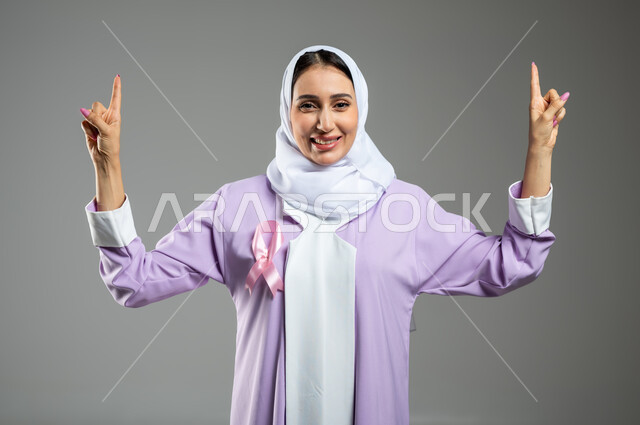 شعار اليوم العالمي للتوعيه بالكشف المبكر لسرطان الثدي، الحملة التوعويه بالامراض الخبيثه، بورتريه مقرب لامرأة عربية خليجية سعودية مبتسمة محجبة ترتدي العباءة مع الشريط الوردي تشير بإصبعي السبابة نحو الاعلى للاشارة الى شئ ما، خلفية رمادية، محتوى حساس، استخدام حساس