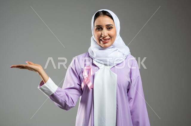 شعار اليوم العالمي للتوعيه بالكشف المبكر لسرطان الثدي، الحملة التوعويه بالامراض الخبيثه، بورتريه لامرأة عربية خليجية سعودية مبتسمة محجبة ترتدي العباءة مع الشريط الوردي تشير بالكف الى شيء ما او لحمل عنصر ما، خلفية رمادية، محتوى حساس، استخدام حساس