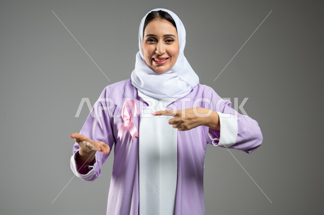 شعار اليوم العالمي للتوعيه بالكشف المبكر لسرطان الثدي، الحملة التوعويه بالامراض الخبيثه، بورتريه لامرأة عربية خليجية سعودية مبتسمة محجبة ترتدي العباءة مع الشريط الوردي تشير بإصبع السبابة نحو اليد الثانية لتحديد شيء معين، خلفية رماديه، محتوى حساس، استخدام حساس