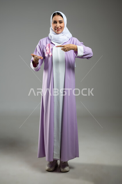 شعار اليوم العالمي للتوعيه بالكشف المبكر لسرطان الثدي، الحملة التوعويه بالامراض الخبيثه، بورتريه لامرأة عربية خليجية سعودية مبتسمة محجبة ترتدي العباءة مع الشريط الوردي تشير بإصبع السبابة نحو اليد الثانية لتحديد شيء معين، صورة الجسم بالطول الكامل، خلفية رماديه، محتوى حساس، استخدام حساس