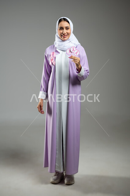 الحملة التوعويه بالامراض الخبيثه، بورتريه مقرب لشابة عربية خليجية سعودية مبتسمة محجبة ترتدي العباءة مع الشريط الوردي تحمل بيدها  شعار التوعيه بالكشف المبكر لسرطان الثدي، صورة الجسم بالطول الكامل، خلفية رماديه، محتوى حساس، استخدام حساس