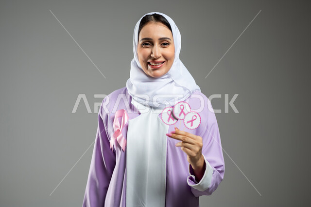 الحملة التوعويه بالامراض الخبيثه، بورتريه مقرب لشابة عربية خليجية سعودية مبتسمة محجبة ترتدي العباءة مع الشريط الوردي تحمل شعار التوعية بالكشف المبكر لسرطان الثدي، خلفية رماديه، محتوى حساس، استخدام حساس