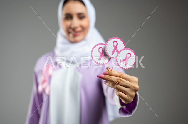 الحملة التوعويه بالامراض الخبيثه، بورتريه مقرب لشابة عربية خليجية سعودية مبتسمة محجبة ترتدي العباءة مع الشريط الوردي تحمل بيدها شعار التوعية بالكشف المبكر لسرطان الثدي، خلفية رماديه، محتوى حساس، استخدام حساس