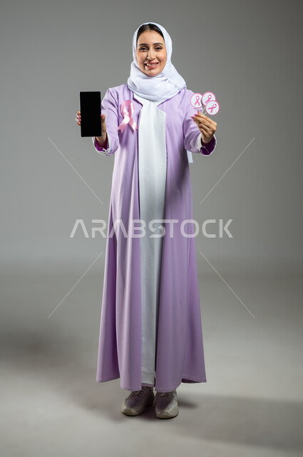 Looking at the camera with expressions of joy and pleasure, a portrait of a smiling Saudi Gulf Arab young woman, veiled, wearing an abaya, displaying a blank, black screen on a mobile phone and holding the Breast Cancer Awareness logo in her other hand, full-length body photo, gray background, sensitive content, sensitive use