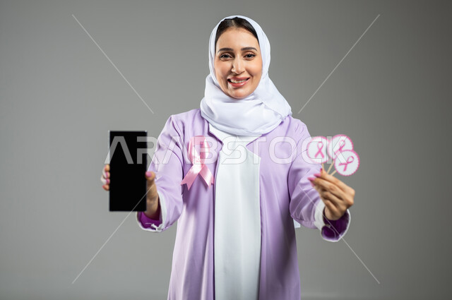 الحملة التوعويه بالامراض الخبيثه، بورتريه مقرب لشابة عربية خليجية سعودية مبتسمة محجبة ترتدي العباءة مع الشريط الوردي تقوم بعرض شاشة فارغة سوداءعبر الهاتف المحمول وتحمل بيدها الثانية شعار التوعية بالكشف المبكر لسرطان الثدي، خلفية رماديه، محتوى حساس، استخدام حساس