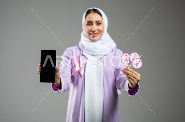 الحملة التوعويه بالامراض الخبيثه، بورتريه لشابة عربية خليجية سعودية مبتسمة محجبة ترتدي العباءة مع الشريط الوردي تقوم بعرض شاشة فارغة سوداءعبر الهاتف المحمول وتحمل بيدها الثانية شعار التوعية بالكشف المبكر لسرطان الثدي،  خلفية رماديه، محتوى حساس، استخدام حساس