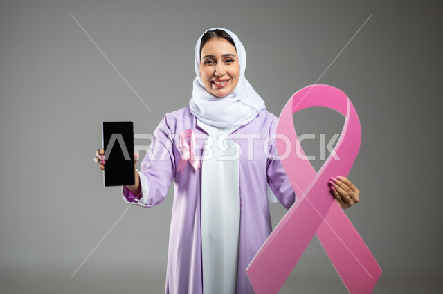 شعار اليوم العالمي للتوعيه بالكشف المبكر لسرطان الثدي، الحملة التوعويه بالامراض الخبيثه، بورتريه لشابة عربية خليجية سعودية مبتسمة محجبة ترتدي العباءة مع الشريط الوردي تقوم بعرض شاشة فارغة سوداءعبر الهاتف المحمول، خلفية رماديه، محتوى حساس، استخدام حساس