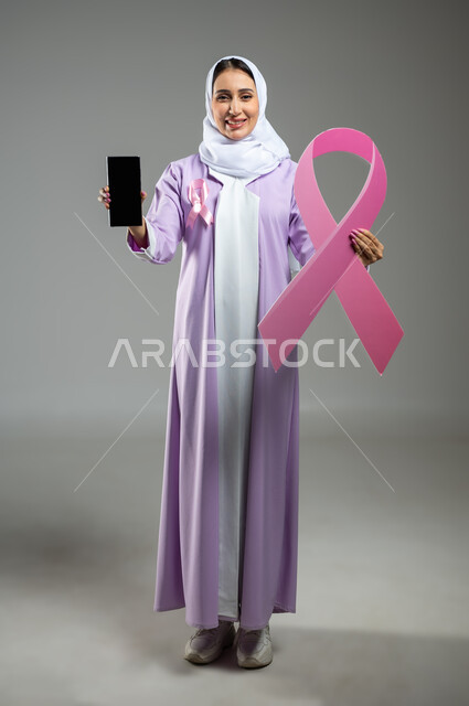 شعار اليوم العالمي للتوعيه بالكشف المبكر لسرطان الثدي، الحملة التوعويه بالامراض الخبيثه، بورتريه لشابة عربية خليجية سعودية مبتسمة محجبة ترتدي العباءة مع الشريط الوردي تقوم بعرض شاشة فارغة سوداءعبر الهاتف المحمول، صورة الجسم بالطول الكامل، خلفية رماديه، محتوى حساس، استخدام حساس