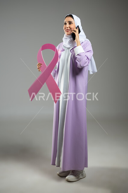 شعار اليوم العالمي للتوعيه بالكشف المبكر لسرطان الثدي، الحملة التوعويه بالامراض الخبيثه، بورتريه لامرأة عربية خليجية سعودية مبتسمة محجبة ترتدي العباءه مع الشريط الوردي تحمل شعار التوعية بالكشف المبكر لسرطان الثدي تتكلم عبر الهاتف اامحمول، صورة الجسم بالطول الكامل، خلفية رماديه، محتوى حساس، استخدام حساس