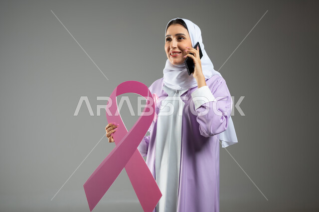 الحملة التوعويه بالامراض الخبيثه، بورتريه لامرأة عربية خليجية سعودية مبتسمة محجبة ترتدي العباءه مع الشريط الوردي  تحمل شعار التوعية بالكشف المبكر لسرطان الثدي تتكلم عبر الهاتف المحمول، خلفية رماديه، محتوى حساس، استخدام حساس