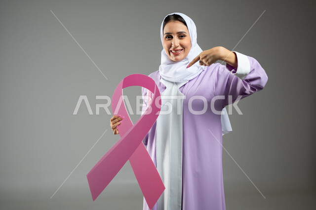 الحملة التوعويه بالامراض الخبيثه، بورتريه لامرأة عربية خليجية سعودية محجبة ترتدي العباءه مع الشريط الوردي تشير بإصبع السبابة نحو  شعار التوعية بالكشف المبكر لسرطان الثدي، خلفية رماديه، محتوى حساس، استخدام حساس