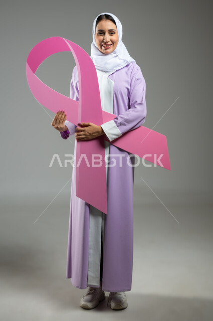 الحملة التوعية بالامراض الخبيثة, بورتريه لإمرأة عربية خليجية سعودية محجبة مبتسمة ترتدي العباءة الملونة تمسك بيدها شعار التوعيه بالكشف المبكر لسرطان الثدي, صورة الجسم بالطول الكامل, خلفية رمادية, محتوى حساس, استخدام حساس