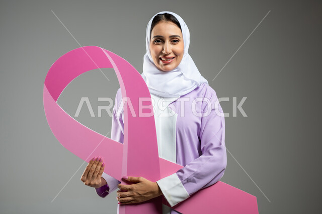 الحملة التوعيه بالامراض الخبيثة, بورتريه مقرب لشابة عربية خليجية سعودية محجبة مبتسمة ترتدي العباءة الملونة تمسك بيدها شعار التوعيه بالكشف المبكر لسرطان الثدي, خلفية رمادية, محتوى حساس, استخدام حساس
