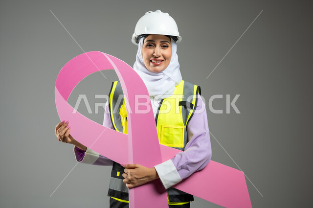 تحدي الأمراض الخبيثة, بورتريه مقرب لإمرأة عربية خليجية سعودية مبتسمة ترتدي سترة الحمايه مع الخوذه تمسك بيديها شعار التوعيه بالكشف المبكر لسرطان الثدي, خلفية رمادية, محتوى حساس, استخدام حساس