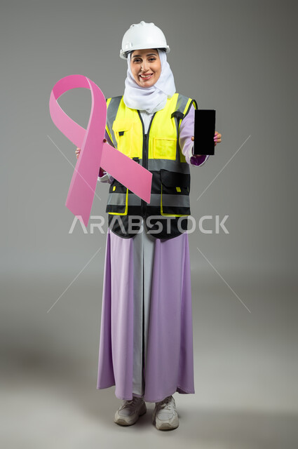 تحدي الأمراض الخبيثة, بورتريه لإمرأة عربية خليجية سعودية مبتسمة ترتدي سترة الحمايه مع الخوذه تمسك بيدها الهاتف المحمول بشاشة فارغة سوداء وشعار التوعيه بالكشف المبكر لسرطان الثدي, صورة الجسم بالطول الكامل, خلفية رمادية, محتوى حساس, استخدام حساس