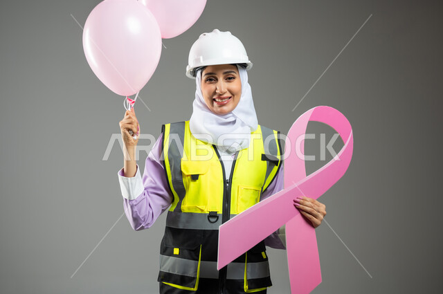 الحملة التوعويه بالامراض الخبيثه، بورتريه مقرب لإمرأة عربية خليجية سعودية مبتسمة محجبة ترتدي سترة الحمايه مع الخوذه تمسك بيدها البلالين الوردية و شعار التوعيه بالكشف المبكر لسرطان الثدي، خلفية رمادية, محتوى حساس, استخدام حساس