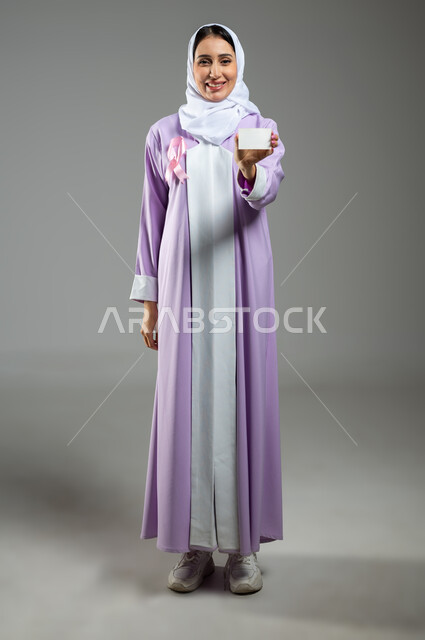 شعار اليوم العالمي للتوعيه بالكشف المبكر لسرطان الثدي، الحملة التوعويه بالامراض الخبيثه، بورتريه مقرب لامرأة عربية خليجية سعودية مبتسمة محجبة ترتدي العباءة مع الشريط الوردي  تحمل بيدها كرت خاص أبيض، خلفية رماديه، صورة الجسم بالطول الكامل، محتوى حساس، استخدام حساس