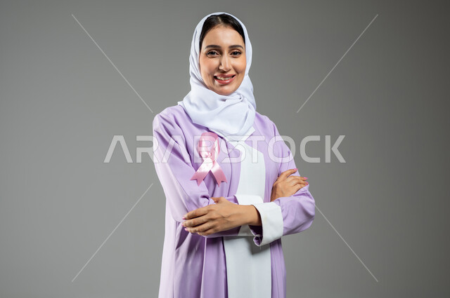Looking at the camera with an expression of pleasure, the logo of the Global Campaign to Challenge Malignant Diseases, a close-up portrait of a smiling, veiled Saudi Arabian Gulf woman wearing an abaya, standing straight with crossed hand gestures, gray background, sensitive content, sensitive use
