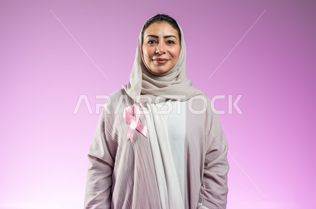 شعار اليوم العالمي للتوعيه بالكشف المبكر لسرطان الثدي، الحملة التوعويه بالامراض الخبيثه، بورتريه مقرب لشابة عربية خليجية سعودية مبتسمة محجبة ترتدي عباءة مع الشريط الوردي تقف باستقامة، خلفية ورديه، محتوى حساس، استخدام حساس