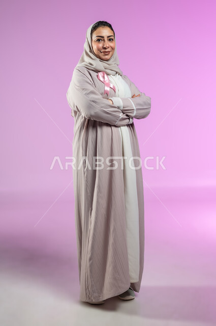 شعار اليوم العالمي للتوعيه بالكشف المبكر لسرطان الثدي، الحملة التوعويه بالامراض الخبيثه، بورتريه مقرب لامرأة عربية خليجية سعودية مبتسمة محجبة ترتدي العباءة مع الشريط الوردي تقف بايماءات ايدي متقاطعة، صورة الجسم بالطول الكامل، خلفية ورديه، محتوى حساس، استخدام حساس