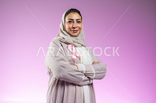 شعار اليوم العالمي للتوعيه بالكشف المبكر لسرطان الثدي، الحملة التوعويه بالامراض الخبيثه، بورتريه مقرب لامرأة عربية خليجية سعودية مبتسمة محجبة ترتدي العباءة مع الشريط الوردي تقف بايماءات ايدي متقاطعة، خلفية ورديه، محتوى حساس، استخدام حساس