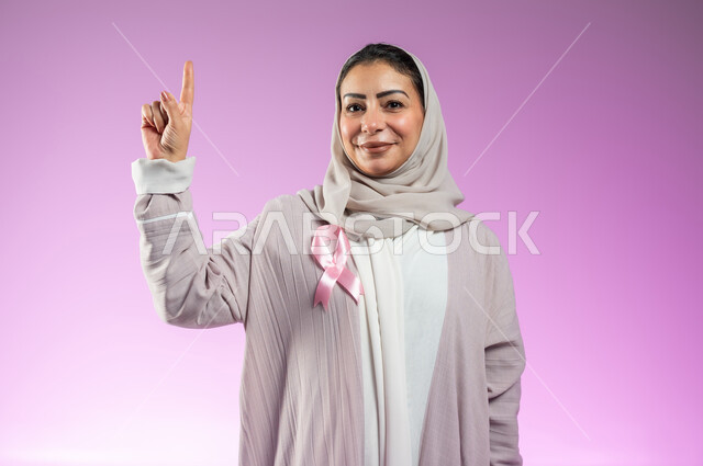 شعار اليوم العالمي للتوعيه بالكشف المبكر لسرطان الثدي، الحملة التوعويه بالامراض الخبيثه، بورتريه مقرب لامرأة عربية خليجية سعودية مبتسمة محجبة ترتدي العباءة مع الشريط الوردي تشير بإصبع السبابة إلى شيء ما في الاعلى، خلفية ورديه، محتوى حساس، استخدام حساس
