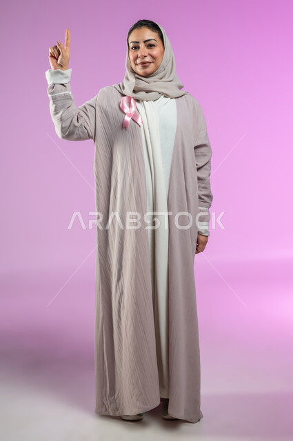 شعار اليوم العالمي للتوعيه بالكشف المبكر لسرطان الثدي، الحملة التوعويه بالامراض الخبيثه، بورتريه مقرب لامرأة عربية خليجية سعودية مبتسمة محجبة ترتدي العباءة مع الشريط الوردي تشير بإصبع السبابة إلى شيء ما في الاعلى، صورة الجسم بالطول الكامل، خلفية ورديه، محتوى حساس، استخدام حساس