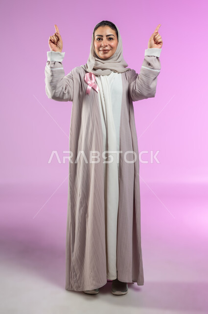 شعار اليوم العالمي للتوعيه بالكشف المبكر لسرطان الثدي، الحملة التوعويه بالامراض الخبيثه، بورتريه مقرب لامرأة عربية خليجية سعودية مبتسمة محجبة ترتدي العباءة مع الشريط الوردي تشير بإصبعي السبابة إلى شيء ما في الاعلى، صورة الجسم بالطول الكامل، خلفية ورديه، محتوى حساس، استخدام حساس