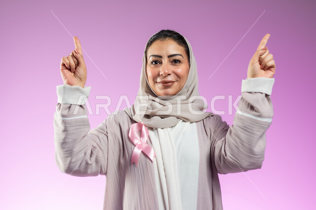 شعار اليوم العالمي للتوعيه بالكشف المبكر لسرطان الثدي، الحملة التوعويه بالامراض الخبيثه، بورتريه مقرب لامرأة عربية خليجية سعودية مبتسمة محجبة ترتدي العباءة مع الشريط الوردي تشير بإصبعي السبابة إلى شيء ما في الأعلى، خلفية ورديه، محتوى حساس، استخدام حساس