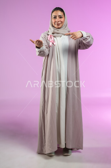 شعار اليوم العالمي للتوعيه بالكشف المبكر لسرطان الثدي، الحملة التوعويه بالامراض الخبيثه، بورتريه لامرأة عربية خليجية سعودية مبتسمة محجبة ترتدي العباءة مع الشريط الوردي تشير بإصبع السبابة نحو اليد الثانية لتحديد شيء معين، صورة الجسم بالطول الكامل، خلفية ورديه، محتوى حساس، استخدام حساس