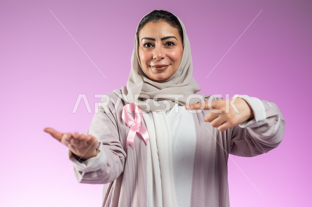 شعار اليوم العالمي للتوعيه بالكشف المبكر لسرطان الثدي، الحملة التوعويه بالامراض الخبيثه، بورتريه لامرأة عربية خليجية سعودية مبتسمة محجبة ترتدي العباءة مع الشريط الوردي تشير بإصبع السبابة نحو اليد الثانية لتحديد شيء معين، خلفية ورديه، محتوى حساس، استخدام حساس