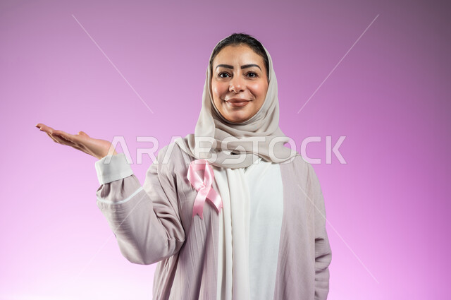 شعار اليوم العالمي للتوعيه بالكشف المبكر لسرطان الثدي، الحملة التوعويه بالامراض الخبيثه، بورتريه لامرأة عربية خليجية سعودية مبتسمة محجبة ترتدي العباءة مع الشريط الوردي تشير بالكف لحمل شيء ما، خلفية ورديه، محتوى حساس، استخدام حساس