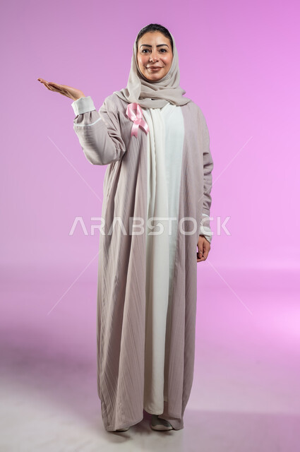 شعار اليوم العالمي للتوعيه بالكشف المبكر لسرطان الثدي، الحملة التوعويه بالامراض الخبيثه، بورتريه لامرأة عربية خليجية سعودية مبتسمة محجبة ترتدي العباءة مع الشريط الوردي تشير بالكف إلى شيء ما و حمل عنصر معين، خلفية ورديه، محتوى حساس، استخدام حساس