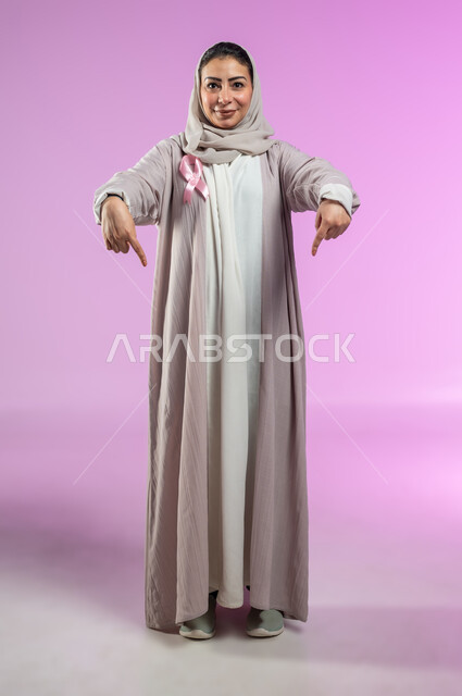 شعار اليوم العالمي للتوعيه بالكشف المبكر لسرطان الثدي، الحملة التوعويه بالامراض الخبيثه، بورتريه لامرأة عربية خليجية سعودية مبتسمة ترتدي الحجاب والعباءة مع الشريط الوردي تشير بإصبعي السبابة نحو الأسفل لتحديد موقع شيء معين، صورة الجسم بالطول الكامل، خلفية ورديه، محتوى حساس، استخدام حساس