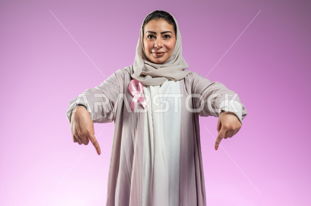 رمز الحملة العالمية، تحدي الأمراض الخبيثة، بورتريه لامرأة عربية خليجية سعودية مبتسمة ترتدي الحجاب و العباءة تشير بإصبعي السبابة نحو الأسفل لتحديد موقع شيء معين، خلفية ورديه، محتوى حساس، استخدام حساس